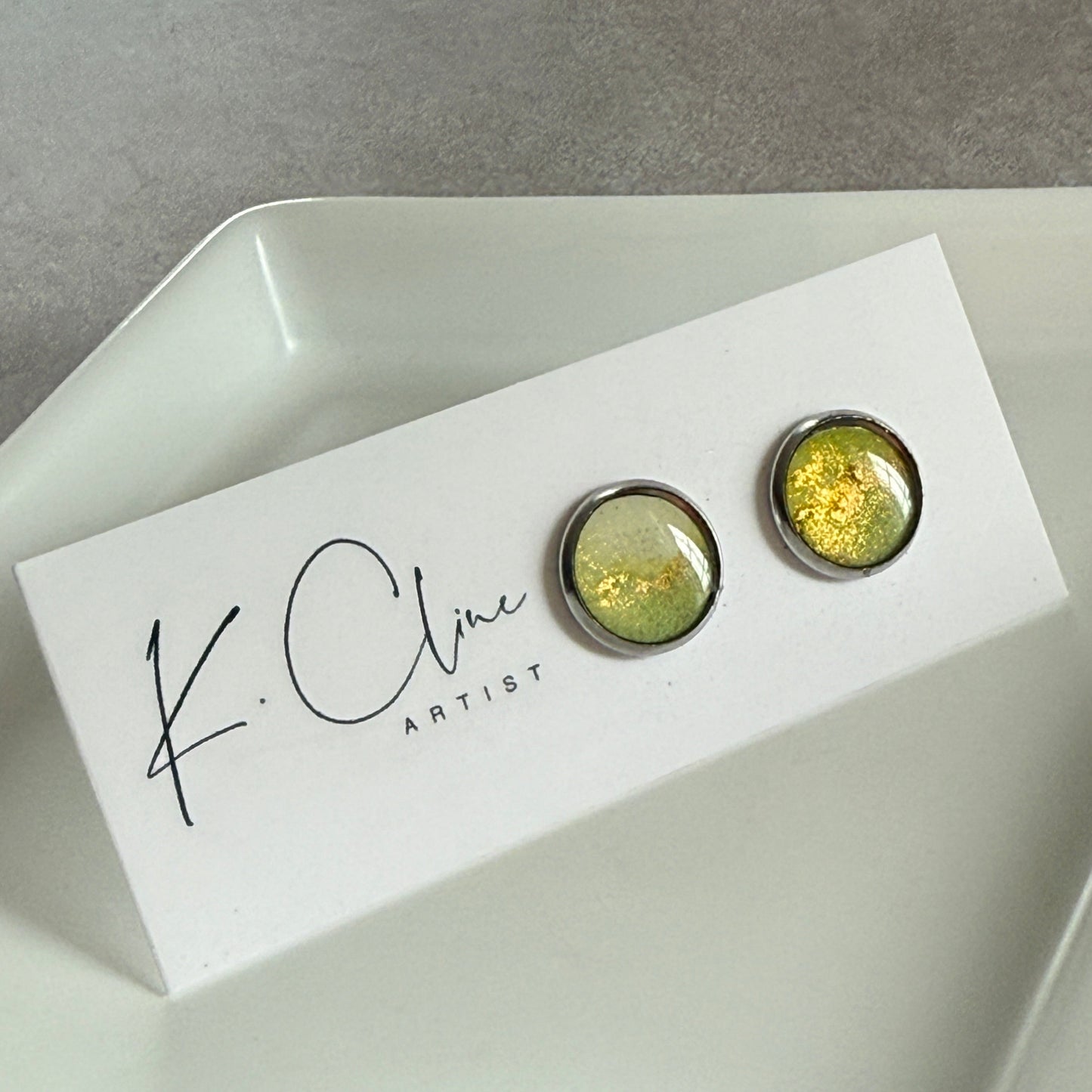 Pair of green earrings on a white box with 'K. Cline Artist' branding on a gray surface.