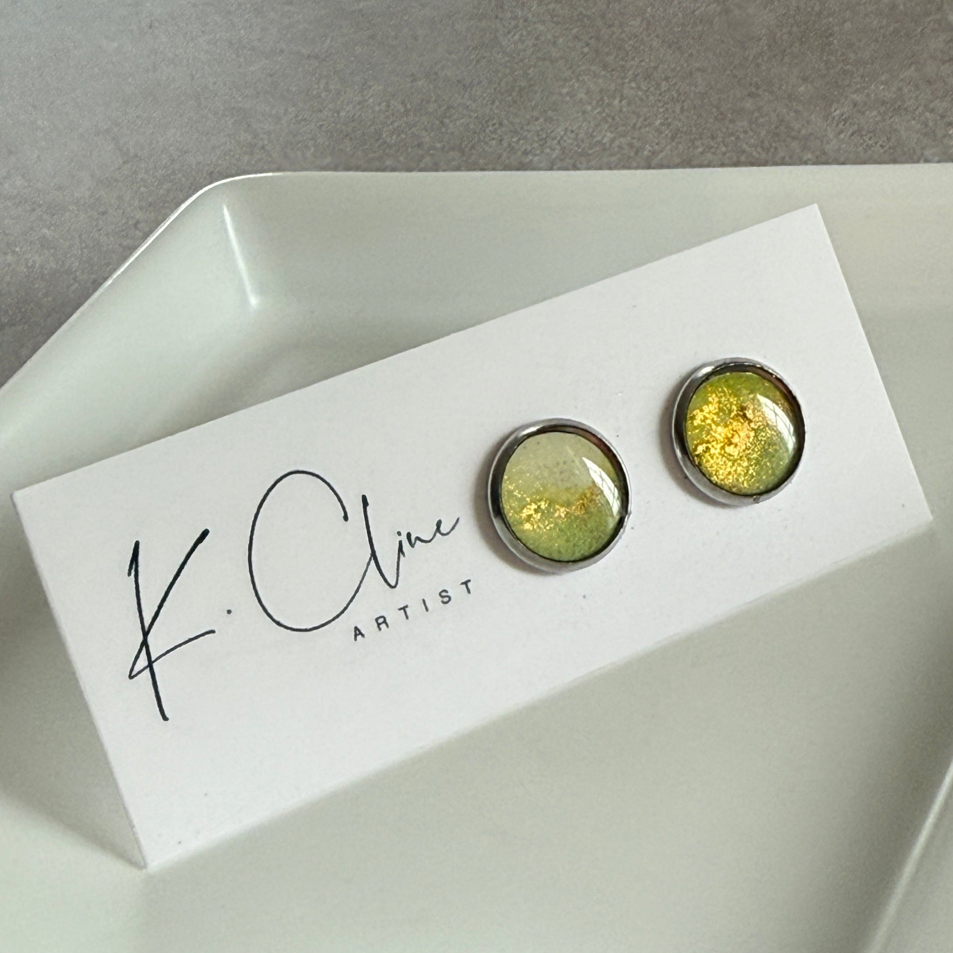 Pair of green earrings on a white box with 'K. Cline Artist' branding on a gray surface.